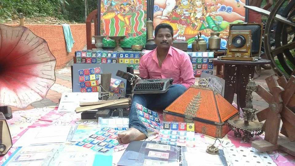 Santhosh seated among his collection — a typewriter, stamp albums, coins, brass lamps, a gramophone horn and a plate camera laid out around him, with Kerala mural paintings behind.